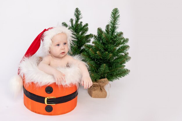 Uma criança pequena está sentada na cesta do Papai Noel em um fundo branco e isolado em um chapéu e com árvores de Natal, o conceito de feliz ano novo e o Natal, um lugar para texto