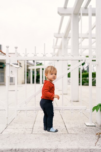 Uma criança pequena fica em pé nas lajes do pavimento e leva a mão a um portão de metal branco fechado