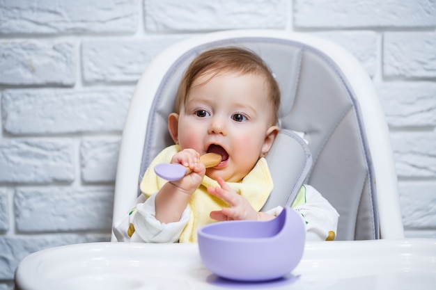 Uma criança pequena se senta em uma cadeira alta e come a comida em um prato com uma colher. Utensílios de silicone para bebês para alimentar bebês