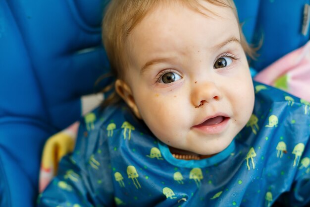 Uma criança pequena se senta em uma cadeira de alimentação e come pão pela primeira vez. Rosto em migalhas