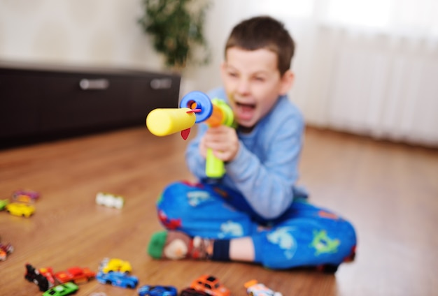 Uma criança pequena, um menino em idade pré-escolar, está jogando um jogo de tiro com uma arma de brinquedo no fundo de uma sala de jogos iluminada com brinquedos. uma bala de espuma ou foguete sai voando de uma metralhadora de brinquedo.