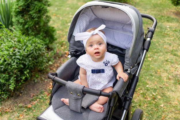 Uma criança pequena, uma menina de 7 meses, senta-se em um carrinho no verão em um vestido branco e olha para a câmera