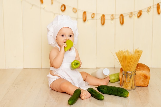 Uma criança pequena, uma menina de seis meses, está sentada na cozinha com um chapéu e um avental de chef e comendo pimentão verde