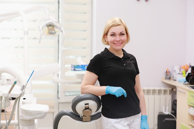 Uma dentista de uniforme posa contra um fundo de equipamentos odontológicos em um consultório odontológico.