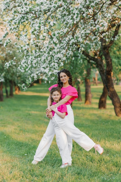 Uma doce jovem de calça branca e blusa rosa brilhante com a filha está caminhando por um parque ou jardim de primavera entre as árvores floridas de uma macieira e a abraça