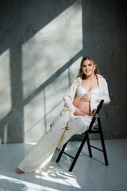 Uma elegante jovem grávida de terno branco senta-se em uma cadeira e toca sua barriga com um sorriso gentil Aguardando o nascimento Cuidados e maternidade Amor e ternura