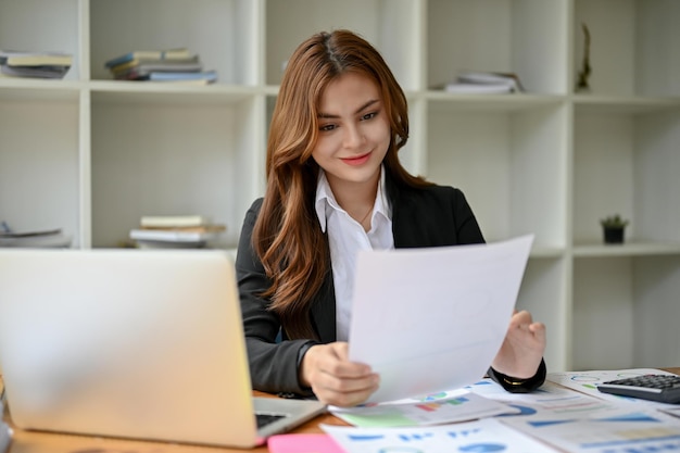 Uma empresária asiática positiva está lendo um relatório de negócios em sua mesa em seu escritório