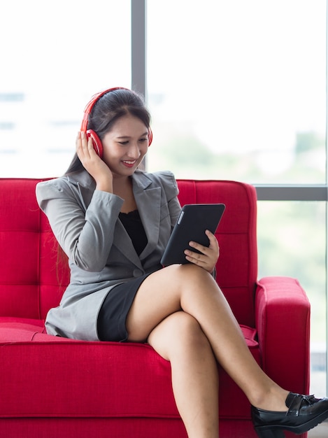 Uma empresária asiática sonhadora usando fones de ouvido, sentada no sofá no corredor do centro de negócios e curtindo músicas com pose fácil durante o intervalo do trabalho