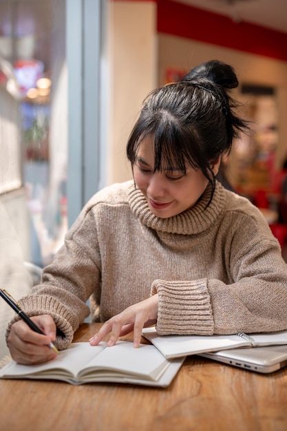 Uma encantadora mulher asiática está listando suas ideias em um livro trabalhando remotamente em uma cafeteria