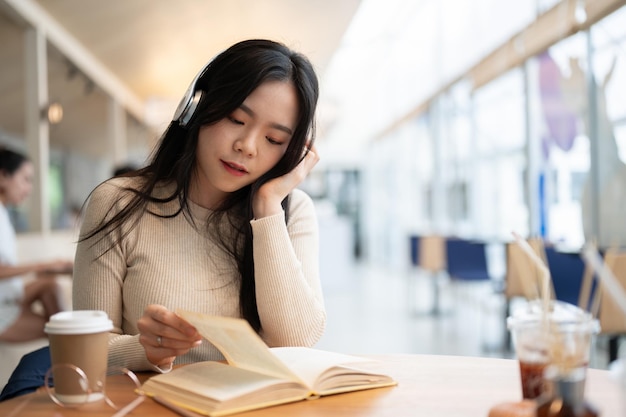 Uma encantadora mulher asiática está ouvindo música através de seus fones de ouvido enquanto lê um livro