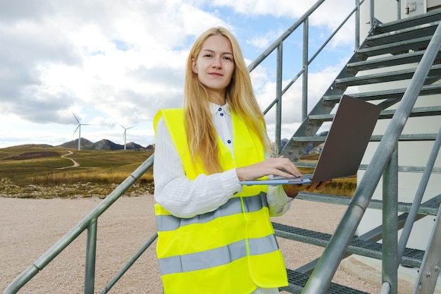 Uma engenheira de energia feminina com um laptop enquanto configura uma turbina eólica Uma jovem está de pé nas escadas de uma turbina eólica