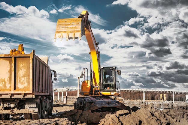 Uma escavadeira carrega um caminhão basculante com solo e areia Uma escavadeira com um balde alto contra um céu nublado Vista da trincheira Remoção de solo de um canteiro de obras ou pedreira