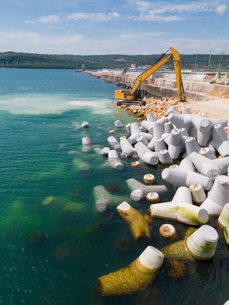 Uma escavadeira constrói diligentemente uma doca ou quebra-mar no mar, seu poderoso braço estendendo-se da costa criando uma estrutura resistente em meio às ondas Vista aérea