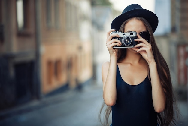Uma esguia menina morena com cabelo comprido, um grande chapéu e uma camiseta preta segura uma câmera de filme antigo nas mãos