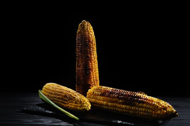 Uma espiga de milho assada sobre carvão em uma placa de madeira espigas de milho inteiras fundo escuro