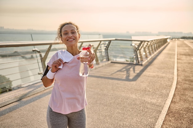 Uma esportista de meia-idade em uma camiseta rosa e legging cinza, com pulseiras brancas, segura uma toalha felpuda nos ombros e sorri alegremente, mostrando uma garrafa de água para a câmera