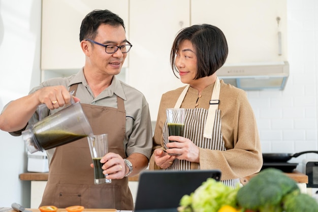 Uma esposa e um marido asiáticos felizes estão desfrutando de um smoothie verde saudável na cozinha juntos