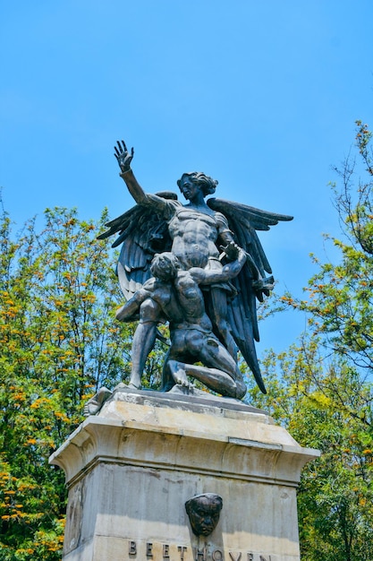 Uma estátua de um homem com asas está em um pedestal com árvores ao fundo