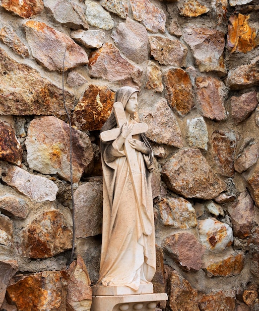 Uma estátua de uma mulher segurando uma cruz fica em frente a uma parede de pedra.