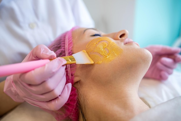 Uma esteticista profissional aplica uma máscara de cura dourada ao rosto de uma mulher Cosmetologia e cuidados com a pele facial em um salão de beleza Procedimento cosmético