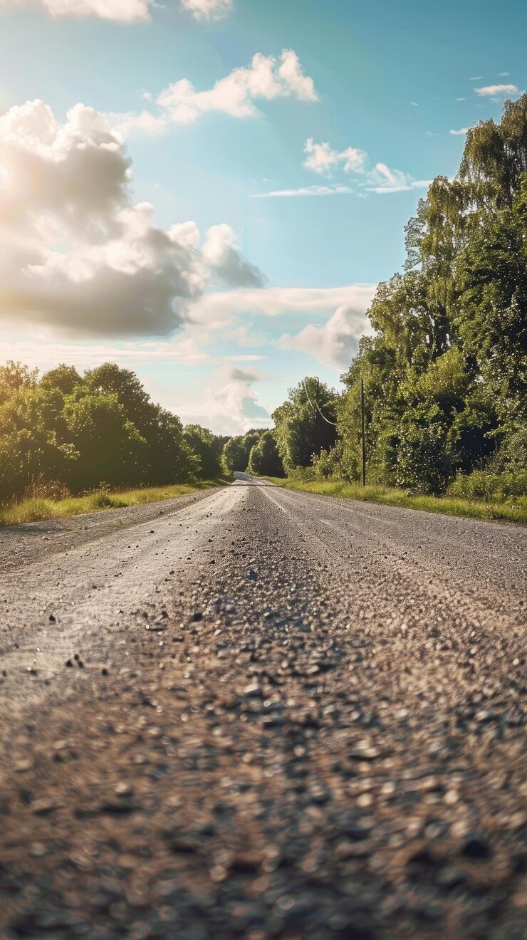 Uma estrada de cascalho se estende à distância emoldurada por árvores e um céu azul com nuvens fofas