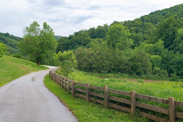 Uma estrada de terra com uma área cercada e um campo gramado com montanhas ao fundo.