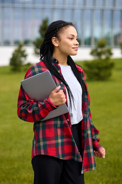 Uma estudante com um laptop no fundo de edifícios modernos Mulher afro-americana estilosa