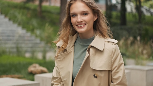 Uma estudante loira bastante positiva vestida com casaco de trincheira parecendo feliz descansando no banco no parque após o estudo