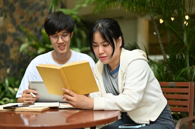 Uma estudante universitária asiática concentrada está lendo um livro enquanto seu amigo está usando um tablet