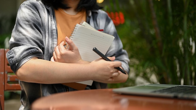 Uma estudante universitária em roupas casuais sentada em um café ao ar livre com seus cozinheiros e laptop