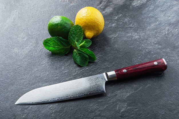 Uma faca japonesa feita de aço damasco repousa sobre uma mesa perto de hortelã e frutas cítricas. vista de cima