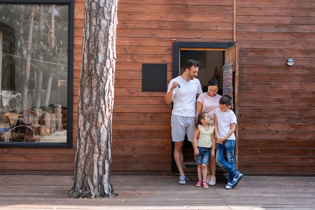 Foto uma família com dois filhos está parada na porta de sua nova casa de madeira fora da cidade, o pai segura as chaves de sua nova casa, todos olham para a menina