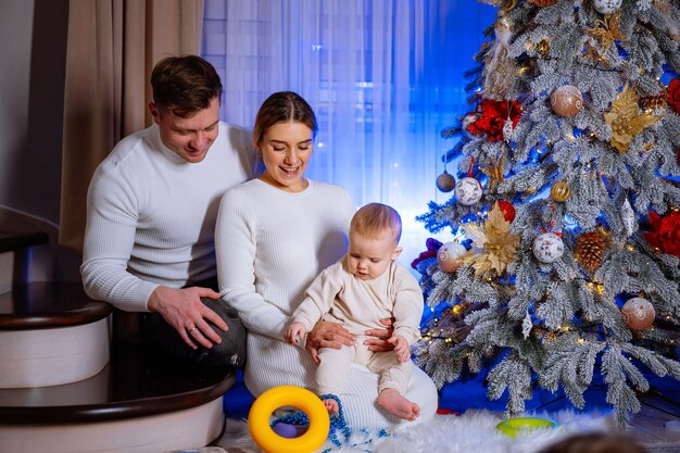 Uma família de três, um homem, uma mulher e um bebê, estão sentados em frente a uma árvore de Natal.