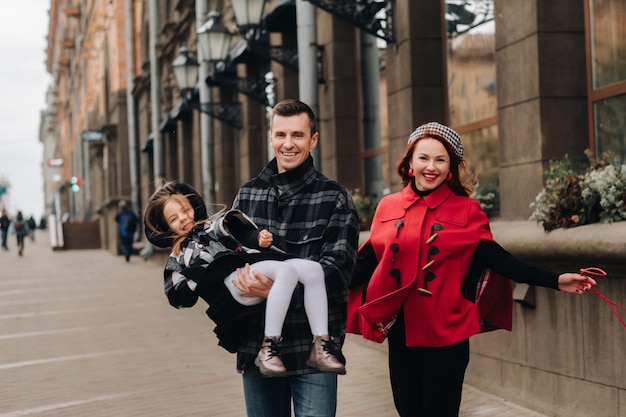 Uma família elegante de três pessoas está andando na cidade no outono e o pai está segurando sua filha nos braços