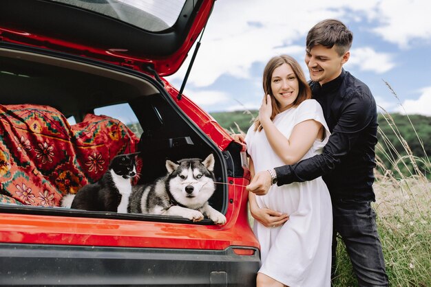 Uma família feliz está esperando um bebê e descansando com um gato e cachorro na natureza perto de seu carro