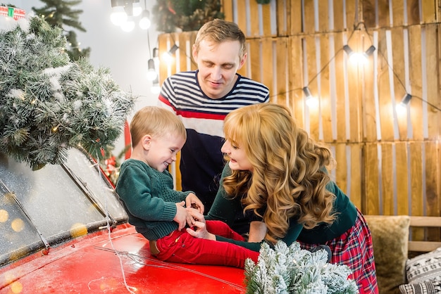 Uma família feliz se divertindo, brincando com o filho perto de um carro vermelho com presentes, árvores de natal