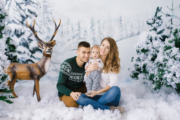 Uma família linda e feliz de jovens pais e um bebê em suéteres de inverno em uma floresta de inverno falsa, na zona de fotos do estúdio