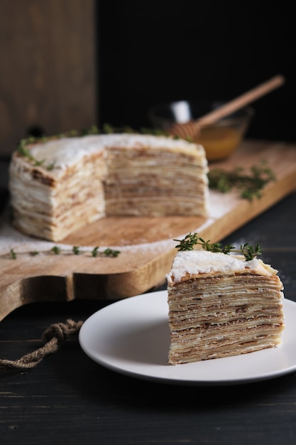 Uma fatia de bolo em um prato. bolo de panqueca com creme de leite, decorado com açúcar de confeiteiro e raminhos de tomilho. sobremesa em uma grande placa de madeira. prato em uma velha mesa de cozinha rústica.