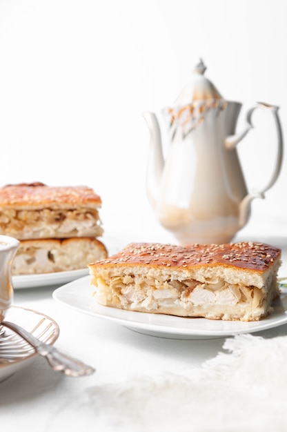 Uma fatia de frango caseiro com torta de gergelim. Num prato branco. Perto de uma cafeteira e uma xícara de café. Fundo branco.