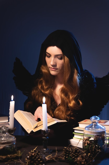 Foto uma feiticeira com uma capa preta com capuz lisonjeia um livro cercado de velas e acessórios de bruxaria