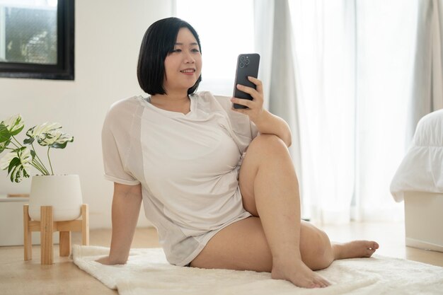 Foto uma feliz mulher asiática plus size em roupas esportivas senta-se em um tapete e usa seu smartphone em seu quarto