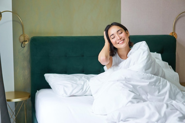 Uma feliz mulher coreana acorda em seu quarto de hotel, deitada na cama sob cobertores quentes e aconchegantes, lençóis brancos enj