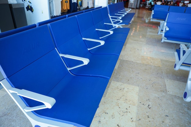 Uma fileira de assentos azuis em uma área de espera com uma planta ao fundo.