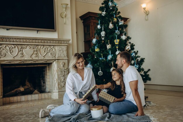 Uma filha abre um presente ao lado de seus pais no fundo de uma árvore de Natal