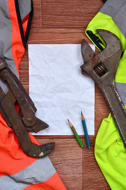 Uma folha de papel amassada com dois lápis rodeados por uniformes de trabalho verdes e laranja e chaves ajustáveis