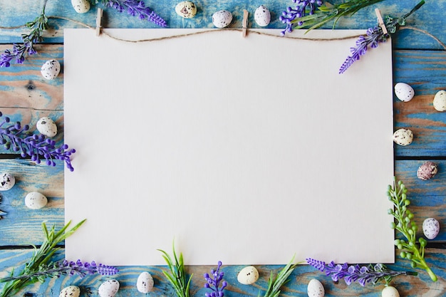 Uma folha de papel branca emoldurada com flores roxas e ovos de codorna em uma mesa de madeira azul gasta