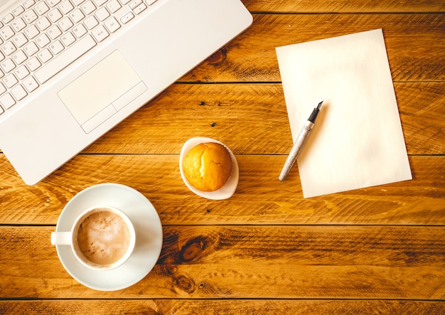 Uma folha de papel em branco com uma caneta sobre uma mesa de madeira com uma xícara de café e um bolo.