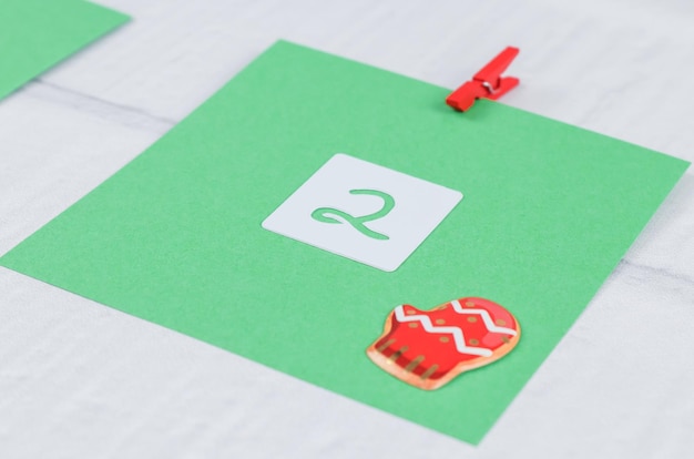 Uma folha de papel verde com números 2 e decoração de Natal anexada com um mini prendedor de roupa vermelho a uma parede de tijolo branco