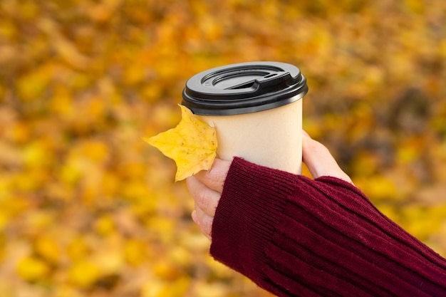 Uma foto aconchegante de uma xícara de café quente artesanal nas mãos contra um fundo de folhas amarelas caídas.