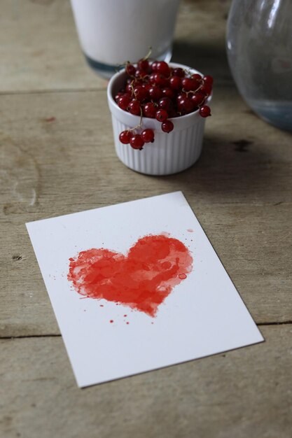 uma foto aproximada de um cartão com um coração aquarela vermelho brilhante em uma mesa de madeira e uma groselha vermelha em